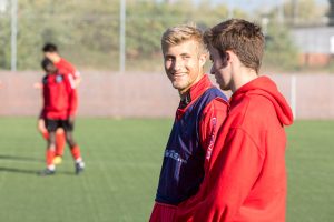 Two young players in red football kit stand on the side of the pitch chatting and laughing 