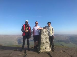 two youths and a man pose by a trig point at the top of a high hill in the UK on a sunny day