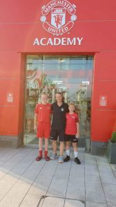 football coach in blue kit standing with two young players outside the Manchester United Youth Academy
