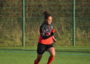 A young female footballer runs to warm up in the sun in her red and black football kit