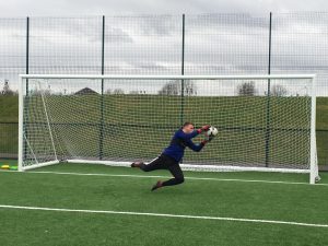 a young goalkeeper in dark blue kit makes a brave dive to save the ball from going into the net