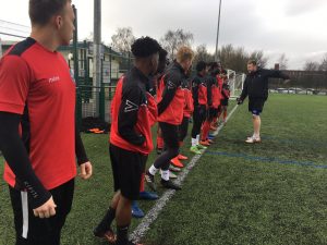 young players dressed in red and black kit line up on the touch line and listen to their coach